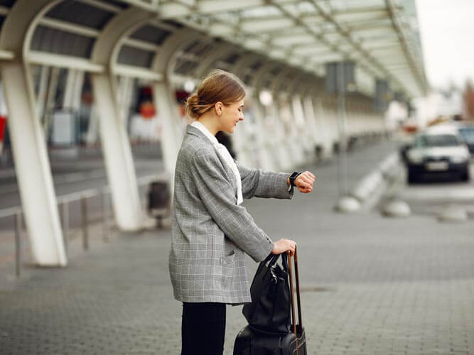 Business travel 2026: ragazza guarda orologio al polso con valigia fuori dall'aeroporto